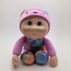 May include: A plush doll with a pink hat and matching outfit. The doll has blue eyes and a smiling expression. The outfit features a rainbow-colored torso and matching rainbow-colored accents on the feet. A blue bow is at the neck.