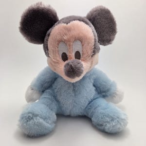 May include: A plush Mickey Mouse toy in a seated position. The toy has a pink face, gray ears, and a gray nose. It is wearing a light blue outfit with white accents on the hands and feet.