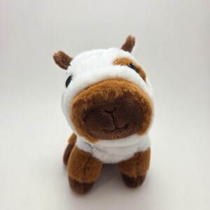 May include: A plush cow toy with a white body, brown spots, and a brown snout. The toy has small brown horns and black eyes. The cow is sitting upright, facing the viewer, and is made of soft, fuzzy material.