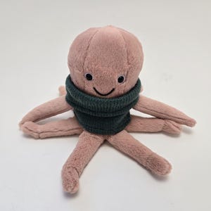 May include: A plush pink octopus wearing a dark green sweater. The octopus has a smiling face with black eyes and a black mouth.