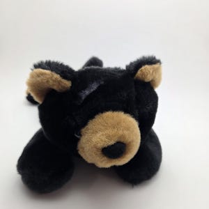 Pode incluir: Um urso de pelúcia preto com detalhes castanhos no focinho e nas orelhas. O bicho de pelúcia está deitado sobre uma superfície branca. O urso tem uma textura macia e peluda.