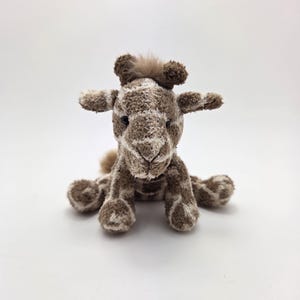 May include: A small, brown and white plush giraffe toy with a fuzzy head and black eyes. The giraffe is sitting on a white background.