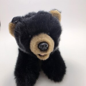 May include: A plush bear toy featuring soft black fur covering the body and head, with contrasting tan fur on the muzzle and inner ears. The bear has dark, round eyes and a smooth, black plastic nose, with its front paws extended forward.