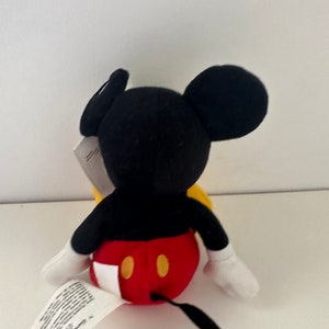 May include: A plush Mickey Mouse toy with black ears, a red and yellow outfit, and a black tail. The toy is sitting on a white surface.