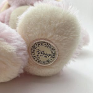 May include: A plush, pink and cream-colored stuffed animal with a Disney Store Authentic tag on its foot. The toy has soft, fluffy fur and a light pink hue, with cream-colored accents on its paws.
