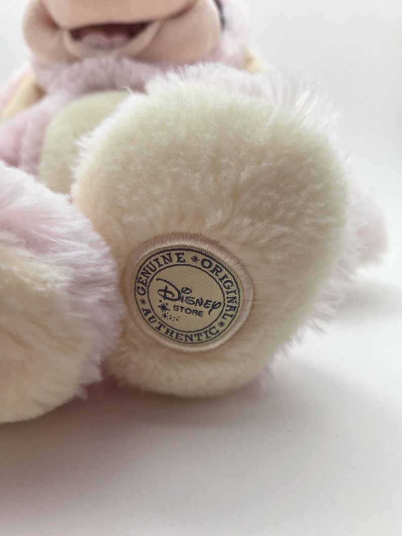 May include: A plush, pink and cream-colored stuffed animal with a Disney Store Authentic tag on its foot. The toy has soft, fluffy fur and a light pink hue, with cream-colored accents on its paws.