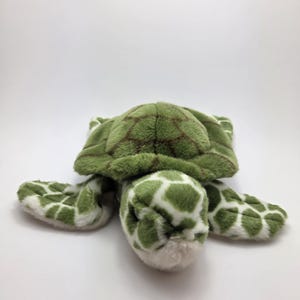 Puede incluir: Un juguete de peluche verde con forma de tortuga marina con caparazón y aletas estampadas. El caparazón de la tortuga tiene un patrón de cuadrícula marrón y sus aletas tienen manchas blancas y verdes. La cabeza y los pies de la tortuga son blancos.