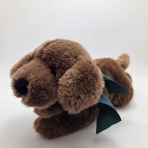 Peut inclure: Un jouet en peluche en forme de chien marron avec un nez noir et des oreilles tombantes. Le jouet a un ruban vert autour du cou. Le chien est allongé sur une surface blanche.