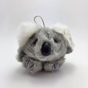 May include: A plush koala toy with gray fur and white accents around the ears. The koala has a black nose and gray paws. A white string is attached to the top, and a small tag is visible. The toy is designed to look like a koala bear.