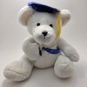 May include: A white teddy bear wearing a blue and yellow graduation cap and a blue ribbon with the letters "SFU". The bear has black eyes and a black nose. The bear is sitting on a white surface.