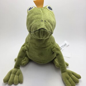 May include: A plush green frog toy with large, round eyes and a small, yellow crown. The frog has outstretched arms and legs with detailed webbed feet. The toy is made of soft, textured fabric.