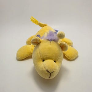 Puede incluir: Un peluche amarillo con forma de animal, posiblemente una jirafa. Tiene una cabeza redonda, orejas pequeñas y un cuerpo largo con cuatro extremidades. El juguete tiene una melena amarilla y un mechón de pelo morado.