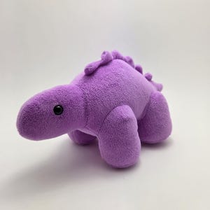 May include: A plush purple dinosaur toy with a rounded body, four stubby legs, and a row of small spikes along its back. The dinosaur has a small head with a black eye on each side. The toy is made of soft, fuzzy material.