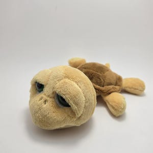 May include: A plush turtle toy with a tan head and a brown shell. The turtle has large, green eyes and soft, fuzzy textures. The toy is lying on a white surface.
