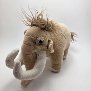 May include: A soft toy mammoth in shades of beige and cream. The stuffed animal has large, curved tusks, small ears, and a tuft of brown hair on its head. The toy is standing on a white surface.