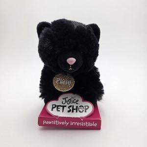 Puede incluir: Un oso de peluche negro con una nariz rosa y una etiqueta dorada que dice "Pixie". El oso está sobre una caja rosa con las palabras "Justice PET SHOP" y "pawsitively irresistible".