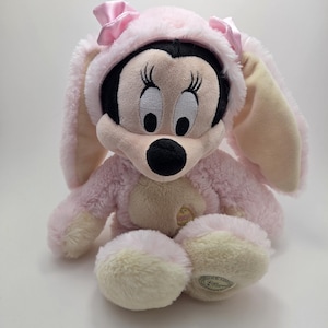 May include: A plush Minnie Mouse doll wearing a pink bunny costume with long ears and a matching hood. The doll has a black face, large eyes, and a pink bow on the hood. The plush toy is sitting on a white surface.