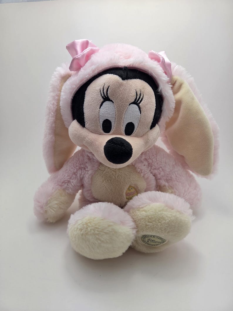 May include: A plush Minnie Mouse doll wearing a pink bunny costume with long ears and a matching hood. The doll has a black face, large eyes, and a pink bow on the hood. The plush toy is sitting on a white surface.