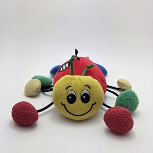 May include: A plush toy shaped like a smiling ladybug. The toy has a yellow head with black eyes and a stitched smile. The body is red with blue wings, and it has six multi-colored, textured legs.
