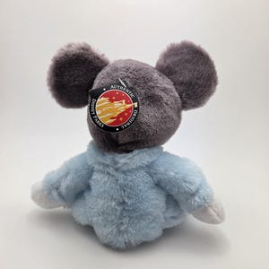 May include: A plush toy mouse with large gray ears and a light blue jacket. The toy has a tag that reads "Disney Parks Authentic Original". The toy is made of soft, fuzzy material.