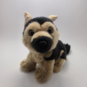 May include: A plush German Shepherd dog toy with black and tan fur, sitting upright. The toy has a black nose, large eyes, and a small tag attached to its chest. The dog's fur is soft and fluffy, and it appears to be new.