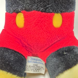 May include: Close-up of a Mickey Mouse plush toy. The toy features a black head and ears, red shorts with two yellow buttons, and yellow feet. A tag reads "Disneyland Walt Disney World".
