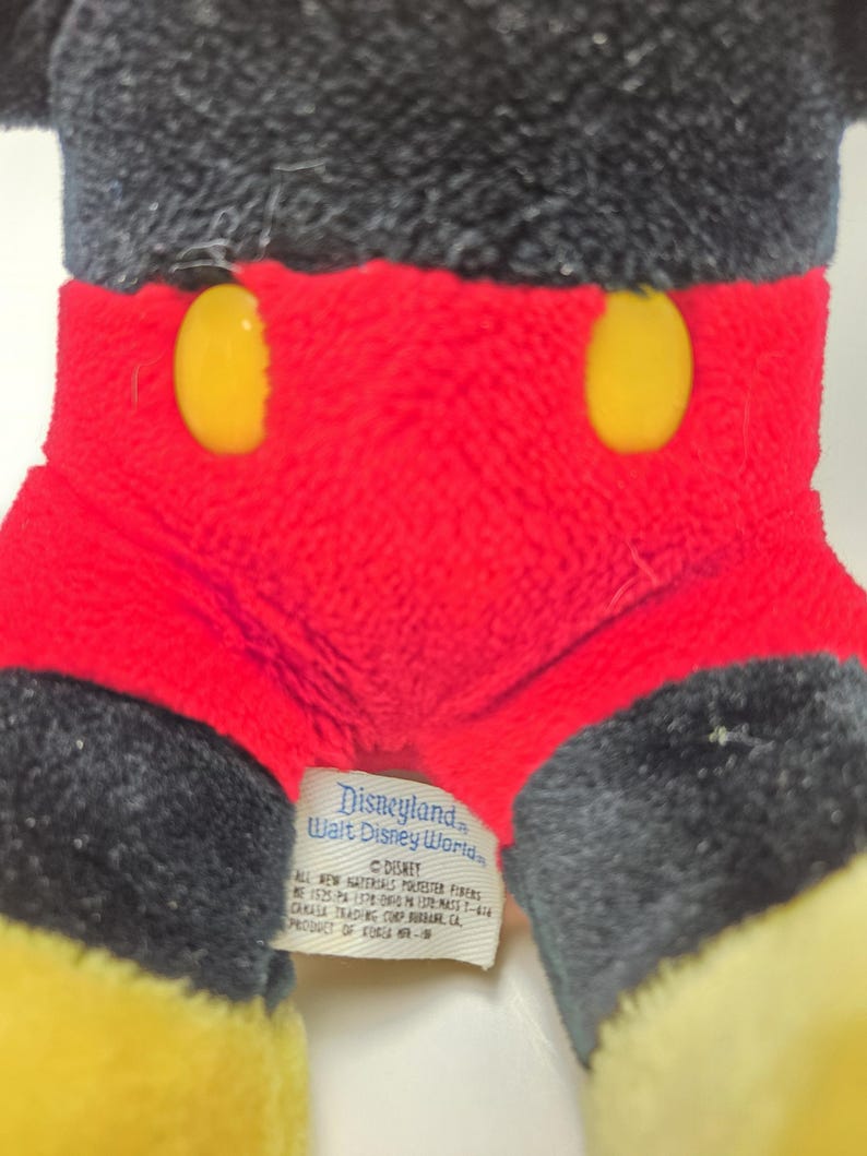 May include: Close-up of a Mickey Mouse plush toy. The toy features a black head and ears, red shorts with two yellow buttons, and yellow feet. A tag reads "Disneyland Walt Disney World".