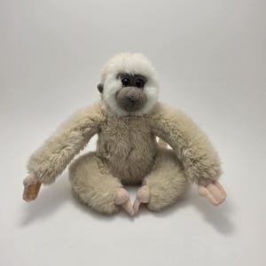 Può includere: Una scimmia di peluche con corpo color crema, testa bianca e viso grigio. Il giocattolo è seduto con le braccia e le gambe distese. Le mani e i piedi della scimmia sono di colore rosa chiaro.