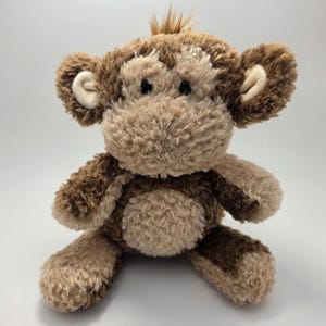 May include: A plush monkey toy with a light brown body and darker brown accents on the ears, hands, and feet. The monkey has a fluffy, textured appearance and a small tuft of brown fur on its head. The toy is designed to sit upright.