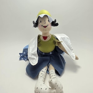 Könnte beinhalten: Handgefertigte Puppe mit bemaltem Gesicht, trägt einen gelben Hut, ein grünes Oberteil und einen blauen Rock. Die Puppe hat weiße Socken mit schwarzen Punkten und einen roten Herzanhänger. Sie hat weiße Stoffflügel und ein blaues sternförmiges Detail.
