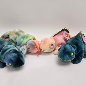 May include: Four colourful plush chameleon beanie babies. The chameleons are green, blue, pink, and tie-dye. Each beanie baby has a red and white tag with a star.