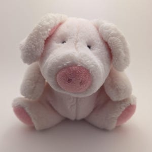 May include: A plush pig toy in a seated position. The pig is primarily white with pink accents on the ears, snout, and feet. It has small black eyes and a soft, cuddly appearance, suitable for children.