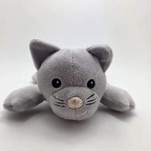Op de afbeelding: Een grijs pluche kattenspeeltje met zwarte ogen, snorharen en een roze neus. De kat ligt met uitgestrekte poten. Het speelgoed is gemaakt van zacht, pluizig materiaal.