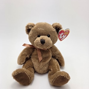 May include: A brown teddy bear with a brown ribbon tied around its neck. The plush toy has a dark brown nose and a red heart-shaped tag attached to its ear. The bear is sitting on a white surface.