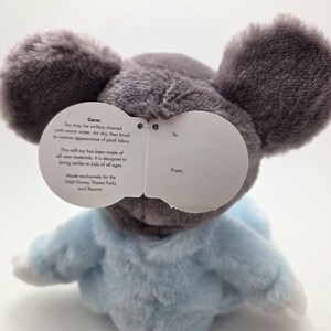 May include: A plush toy mouse with large gray ears and a light blue outfit. The toy has a tag with care instructions and text that reads "Made exclusively for the Walt Disney Theme Parks and Resorts."