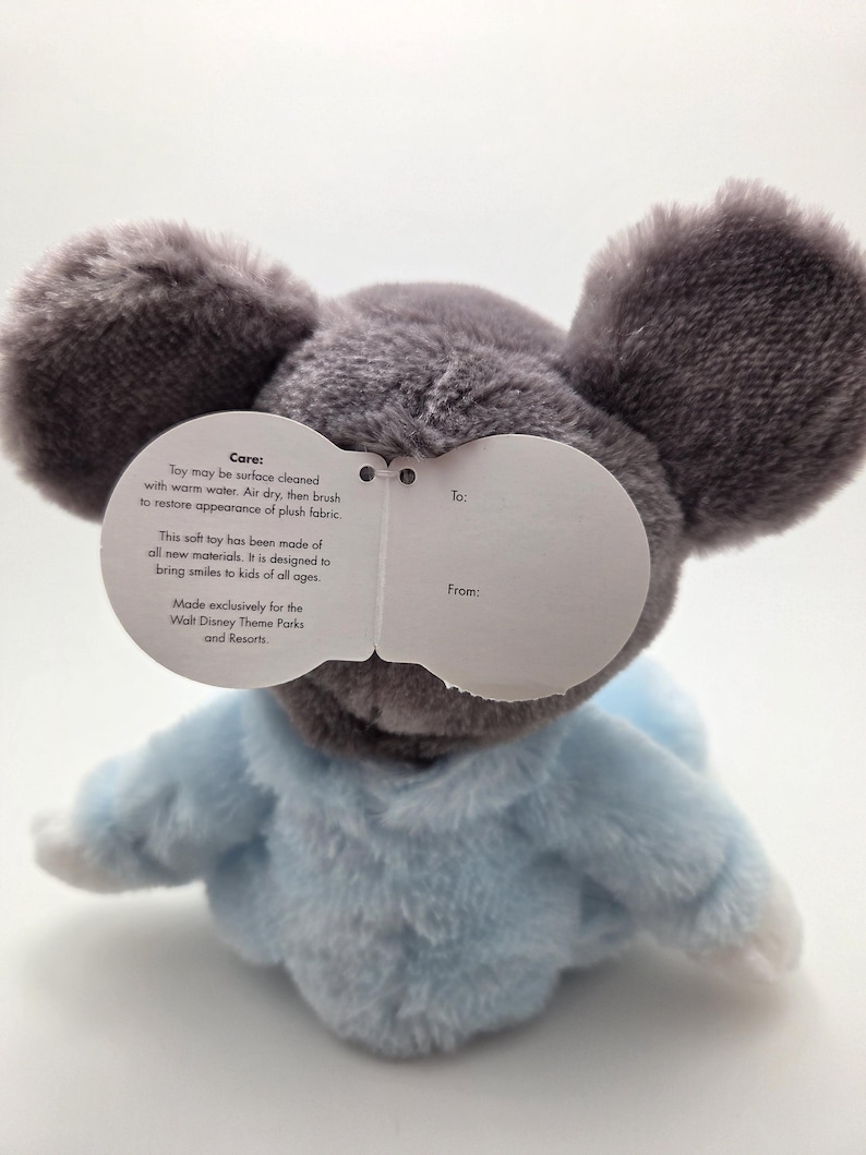 May include: A plush toy mouse with large gray ears and a light blue outfit. The toy has a tag with care instructions and text that reads "Made exclusively for the Walt Disney Theme Parks and Resorts."