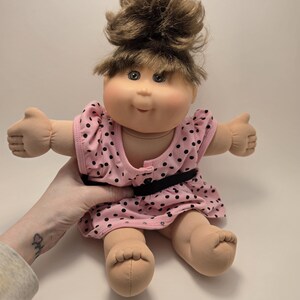 May include: A Cabbage Patch Kids doll with brown hair styled in a bun and bangs. The doll wears a pink dress with black polka dots and a black belt. The doll's arms are outstretched.