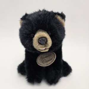 May include: A black plush bear with brown fur around its nose and eyes. The bear is sitting with its paws tucked in front of it. The bear has a brown tag that says "Yomiko Classics" around its neck.