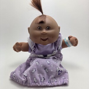 May include: A Cabbage Patch Kids doll with brown skin, brown hair in a topknot, and brown eyes. The doll wears a purple dress with a white trim and a matching headband. A blue and white bracelet is on the doll's wrist.