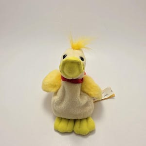 Peut inclure: Un canard en peluche jaune avec un corps crème, des pieds jaunes et un bec jaune. Il porte un ruban rouge autour du cou et une touffe de plumes jaunes sur la tête. Une petite étiquette est attachée.