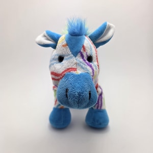 May include: A colorful plush zebra toy with a blue nose, ears, and hooves. The zebra's body is white with rainbow-colored stripes. It has a blue mohawk and black eyes.