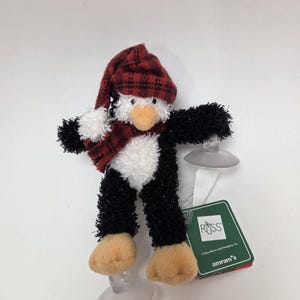 Puede incluir: Un juguete de peluche de pingüino blanco y negro con un gorro y una bufanda de cuadros rojos y negros. El pingüino tiene ventosas en los pies y está unido a una etiqueta verde con el texto "Russ" y "amram's".