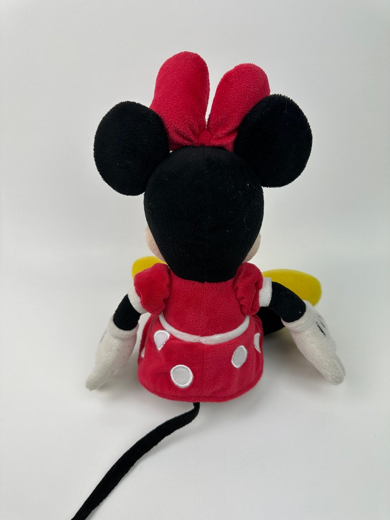 K&ouml;nnte beinhalten: Ein Pl&uuml;schtier von Minnie Mouse, das sitzt. Sie tr&auml;gt ein rotes Kleid mit wei&szlig;en Punkten und eine rote Schleife. Ihre Ohren sind schwarz und ihr Schwanz ist schwarz.