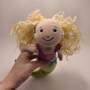 May include: A plush mermaid doll with light skin, a smiling embroidered face, and voluminous blonde yarn hair. The doll wears a purple bodice with yellow lace-up detailing and has a green fabric tail with a subtle scale pattern and blue fringe.