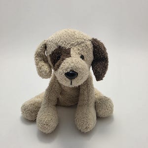 May include: A plush dog toy made of beige textured fabric with a dark brown patch over the right eye and a solid dark brown left ear. The stuffed animal features a smooth black plastic nose and stitched mouth details, sitting in an upright position.
