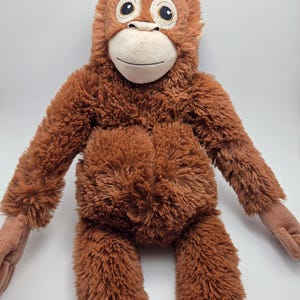 Puede incluir: Un peluche de orangután marrón con una cara de color canela claro y ojos grandes. El peluche tiene brazos y piernas largos, y una expresión sonriente. Un juguete infantil suave y tierno.
