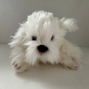 May include: A white plush dog toy with a fluffy, textured coat. The toy has a black nose and small, dark eyes. The dog is lying down, with its paws visible. The overall appearance is soft and cuddly.