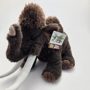 May include: A plush woolly mammoth toy in shades of brown with large white tusks. The toy has a shaggy fur texture and a tag attached to its side. The toy is designed to resemble a prehistoric animal.