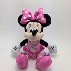 May include: A plush Minnie Mouse doll with a pink and white polka dot bow, black ears, and a pink and white striped top. The doll has pink shoes and a pink tutu. The doll's hands are white with black stripes.