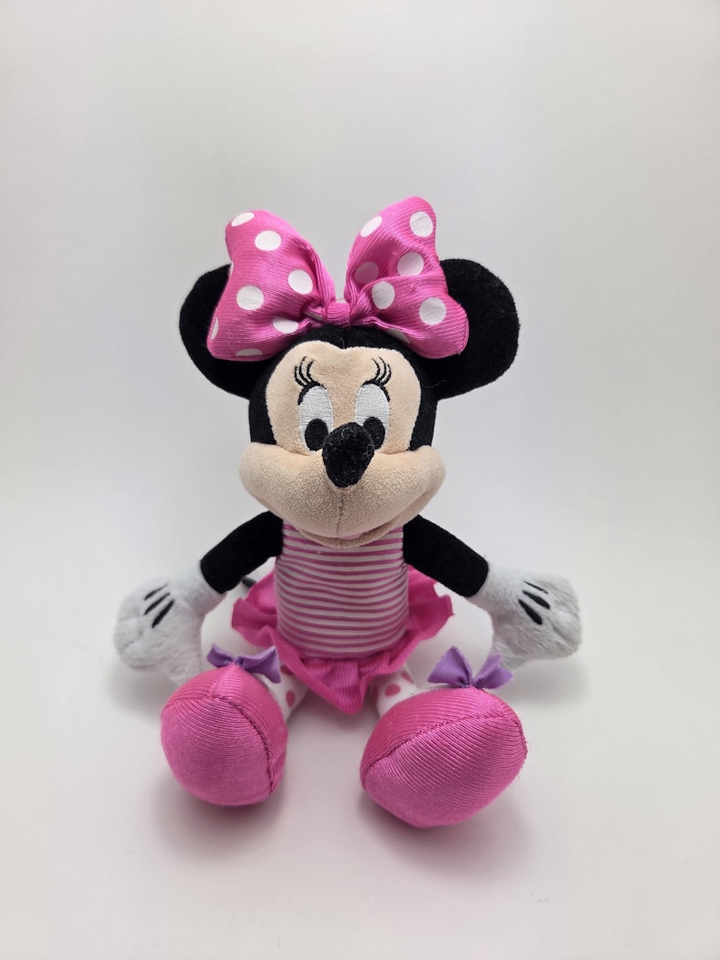 May include: A plush Minnie Mouse doll with a pink and white polka dot bow, black ears, and a pink and white striped top. The doll has pink shoes and a pink tutu. The doll's hands are white with black stripes.